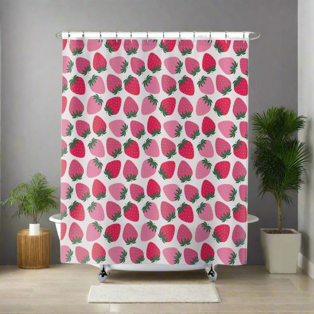 A colorful bathroom shower curtain with a fruit theme, displaying orange and pink fruit patterns against a white background, installed in a bathroom with a plant and a wooden stool in the background.
