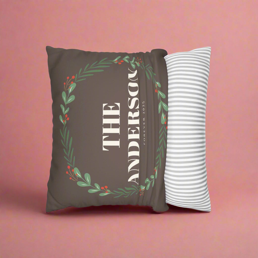 Decorative pillow with 'The Anderson' text and a wreath design on a brown background, next to a white pillow with gray stripes.